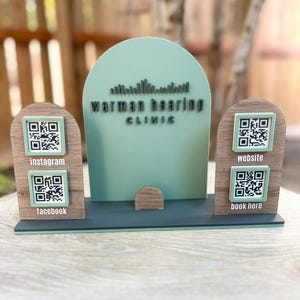 May include: A wooden sign for Warman Hearing Clinic with a mint green arched backdrop and a dark grey base. The sign features QR codes for Instagram, Facebook, website, and booking. The clinic's name is in dark grey.