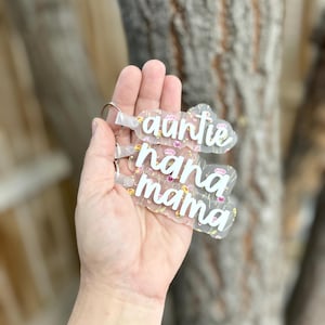 May include: Clear acrylic keyrings with floral patterns and the words "auntie", "nana", and "mama" in white script. Each keyring has a silver ring attached.