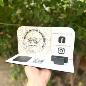 Puede incluir: Un tarjetero blanco y dorado con el texto "HANDCRAFTED WONDERS YXE" e iconos de redes sociales. El soporte tiene una ranura para tarjetas y un código QR. El diseño incluye detalles florales y las iniciales "HW".