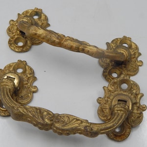 Pode incluir: Duas puxadores de gaveta dourados antigos com desenhos ornamentados de volutas. Cada puxador tem uma alça curva e placas traseiras decorativas com vários orifícios para parafusos. O metal tem uma aparência desgastada, sugerindo idade e uso. Adequado para restauração de móveis antigos.