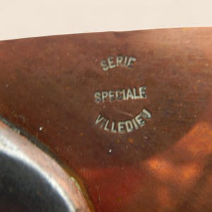 May include: Close-up of a copper-colored metal object with the text "SERIE SPECIALE VILLEDIEJ" in a vintage font. The surface has a slightly reflective quality, and the text appears to be embossed or stamped into the metal.