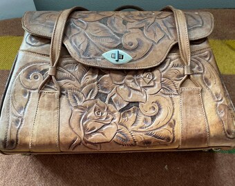 May include: A tan leather handbag featuring an intricate embossed floral pattern across the front flap and body. The bag includes two leather shoulder straps and a central silver-toned metal turn-lock closure mechanism on the front flap.