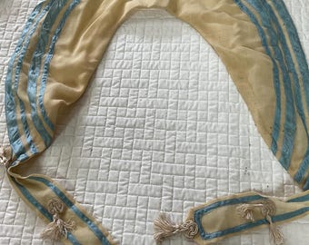 May include: A cream-colored fabric shawl with blue satin trim and tassels. The shawl has a scalloped edge and is tied with a knot at the front.