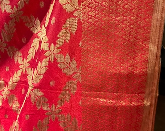 May include: Close-up of a vibrant red fabric with a gold floral pattern. The design features repeating leaf and flower motifs. The fabric has a textured appearance, with a detailed border.