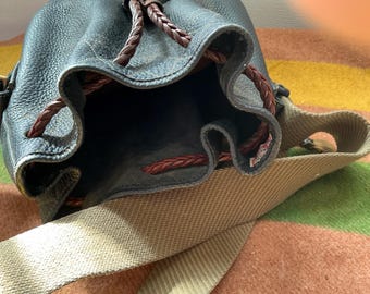 May include: A black leather bucket bag with a textured surface, featuring a dark brown braided drawstring closure and a wide beige canvas shoulder strap. The interior is dark, and the bag has a structured opening with visible stitching details.