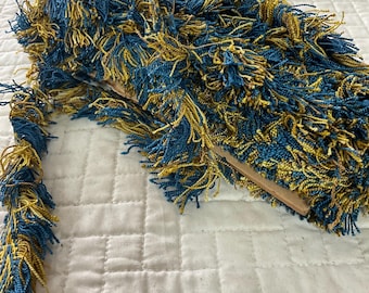 May include: A close-up of a blue and gold fringe trim. The fringe is made of thin strands of yarn that are twisted together. The fringe is about 1 inch wide and is perfect for adding a touch of elegance to any project.