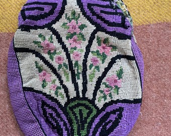 May include: A small, rounded pouch featuring needlepoint embroidery with a floral motif. The design displays a central cream-colored panel with pink flowers and green leaves, framed by bold black outlines and solid purple borders on the sides.