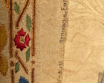 May include: Close-up of a beige fabric with embroidered floral designs in shades of blue, red, green, and yellow. The fabric has a frayed edge with the text "STAMBOUL EMBROIDY" and "8454-91410" printed on it.