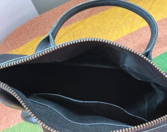May include: A dark blue leather handbag with a top zipper closure featuring a metallic zipper track. The interior is lined with black fabric and includes an open slip pocket along the side wall. The bag has a single rounded handle attached at the top.