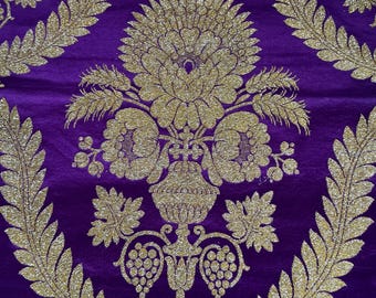 May include: A deep purple fabric featuring a prominent gold metallic floral and urn pattern. The design includes large central blossoms, leafy branches, and grape clusters. A small rectangular tag with handwritten text is attached at the top.