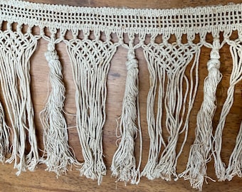 May include: A close-up of a beige cotton fringe trim with a woven pattern. The fringe is made of multiple strands of cotton yarn that are knotted together to create a tassel effect.
