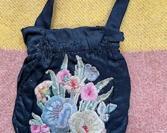 May include: A black satin drawstring pouch featuring a central embroidered floral bouquet. The embroidery includes shades of pink, blue, yellow, and green thread. The bag has a gathered top edge and a single black fabric handle for carrying.