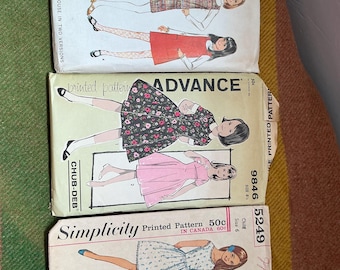 May include: Three vintage sewing pattern envelopes stacked vertically. The top is McCall's 9395 for a girls' jumper and blouse. The middle is Advance 9846 for a dress. The bottom is Simplicity 5249 for a child's dress, each showing illustrations.