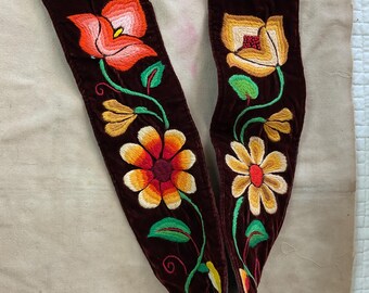 May include: A pair of dark burgundy velvet leg wraps with vibrant floral embroidery. The embroidery features orange, yellow, and white flowers with green leaves and stems. The wraps have a drawstring closure and eyelets.