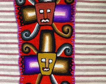 May include: A rectangular, hand-stitched textile with a vibrant red background. Two stylized faces with rectangular bodies and curly accents are featured. The faces are brown and yellow, with purple and brown accents. The edges are finished with purple stitching.