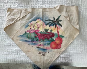 May include: A white cotton bandana with a hand-painted design of pink flamingos, an alligator, palm trees, and oranges. The bandana has the text "Miami Beach Fla." printed on it.
