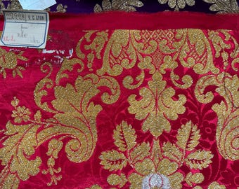 May include: A vibrant red fabric featuring an intricate gold metallic damask pattern with large scrolling acanthus leaves and floral motifs. A small white rectangular paper tag is attached to the top left corner with the text 2228 G.C. LYON.
