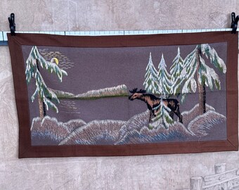 May include: Embroidered wall hanging with a brown background featuring a moose standing in a snowy forest scene with pine trees and a moon.