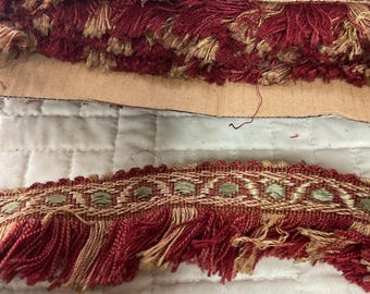 May include: Red and gold fringe trim with a woven pattern. The fringe is made of a soft, textured yarn.