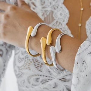 May include: A close-up of a wrist adorned with three open cuff bracelets. Two are gold-toned, and one is silver-toned. The bracelets have a smooth, curved design, with a teardrop-shaped end. The background is a white, lacy fabric.