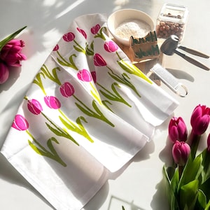 Puede incluir: Toalla de cocina blanca con estampado de tulipanes rosa y verde. La toalla está doblada y sobre una superficie blanca.