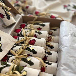 May include: A cardboard box filled with rolled cream-coloured napkins, each adorned with a floral pattern in red, green, and yellow. The napkins are tied with raffia. A matching flat napkin is also visible.