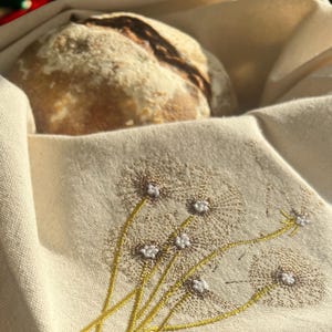 Könnte beinhalten: Ein weißer Leinenstoff mit gestickten Löwenzahnpflanzen in Gold- und Weißtönen. Ein Sauerteigbrot ist im Hintergrund teilweise sichtbar.