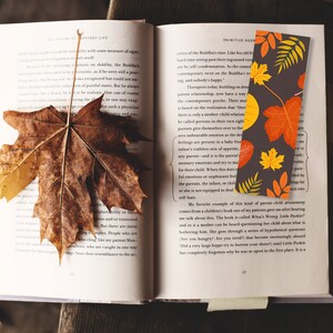 Pode incluir: Um livro aberto com um marcador de página com um design de folhas de outono. O marcador de página está em uma página com o texto "Primitive Agony" e "The Trauma of Everyday Life".