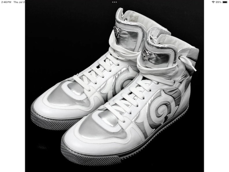 May include: White high-top sneakers with silver accents. The shoes feature a unique design with a textured sole and a lace-up closure. The high-top design includes a padded collar and a decorative emblem.