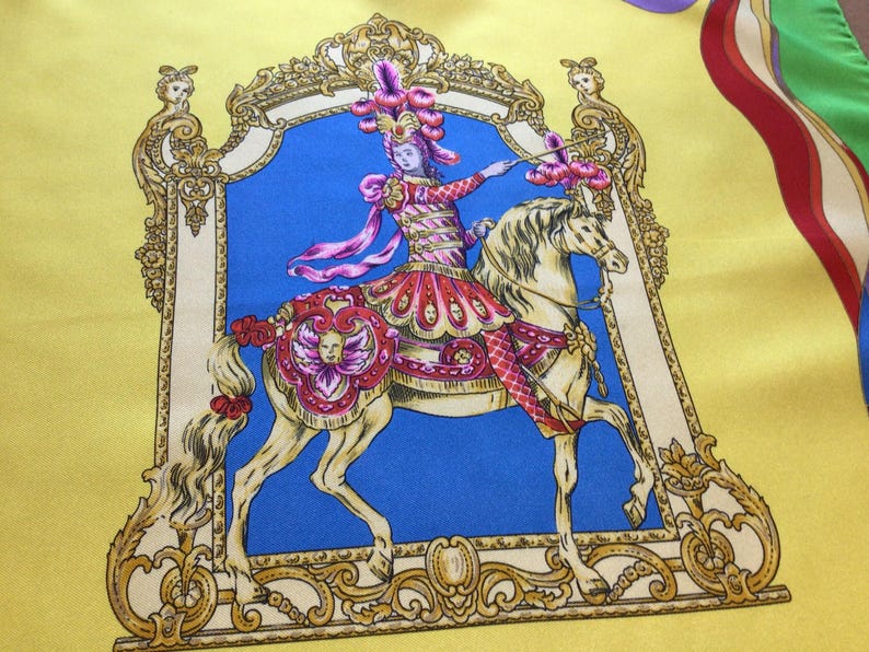 Puede incluir: Pa&ntilde;uelo de seda con una ilustraci&oacute;n detallada de una persona montada a caballo, enmarcada en un borde ornamentado. El dise&ntilde;o presenta un fondo azul, un borde amarillo y detalles en rojo, rosa y dorado.