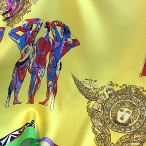 Puede incluir: Un pa&ntilde;uelo de seda amarillo con un dise&ntilde;o vibrante que presenta figuras humanas estilizadas en rojo, azul y morado, junto con notas musicales y elementos decorativos. El pa&ntilde;uelo tambi&eacute;n incluye detalles dorados y bronce ornamentados.