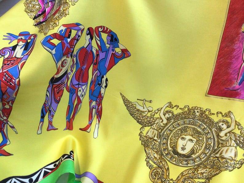 Puede incluir: Un pa&ntilde;uelo de seda amarillo con un dise&ntilde;o vibrante que presenta figuras humanas estilizadas en rojo, azul y morado, junto con notas musicales y elementos decorativos. El pa&ntilde;uelo tambi&eacute;n incluye detalles dorados y bronce ornamentados.