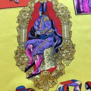 Puede incluir: Pa&ntilde;uelo de seda amarillo con un dise&ntilde;o central de una figura sentada con atuendo morado y rosa, enmarcado por un borde dorado ornamentado. El pa&ntilde;uelo presenta elementos decorativos coloridos adicionales.