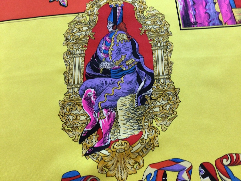 Puede incluir: Pa&ntilde;uelo de seda amarillo con un dise&ntilde;o central de una figura sentada con atuendo morado y rosa, enmarcado por un borde dorado ornamentado. El pa&ntilde;uelo presenta elementos decorativos coloridos adicionales.