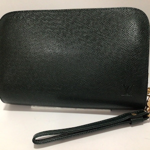 May include: A black rectangular clutch bag with a textured leather finish and rounded corners. The bag features a gold-toned metal clasp and a matching black leather wrist strap attached to the side. A small embossed logo is visible on the bottom right.