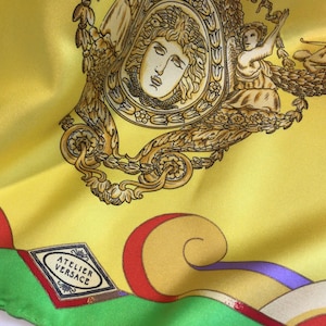 Puede incluir: Pa&ntilde;uelo de seda amarillo con ilustraciones doradas y marrones de figuras cl&aacute;sicas y elementos decorativos. El pa&ntilde;uelo presenta el texto "ATELIER VERSACE" y patrones geom&eacute;tricos coloridos en rojo, verde y azul.