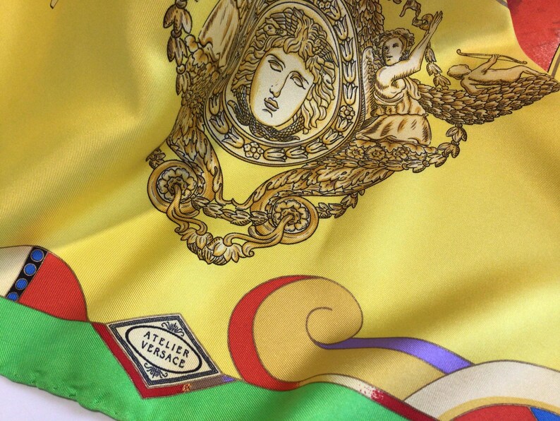 Puede incluir: Pa&ntilde;uelo de seda amarillo con ilustraciones doradas y marrones de figuras cl&aacute;sicas y elementos decorativos. El pa&ntilde;uelo presenta el texto "ATELIER VERSACE" y patrones geom&eacute;tricos coloridos en rojo, verde y azul.