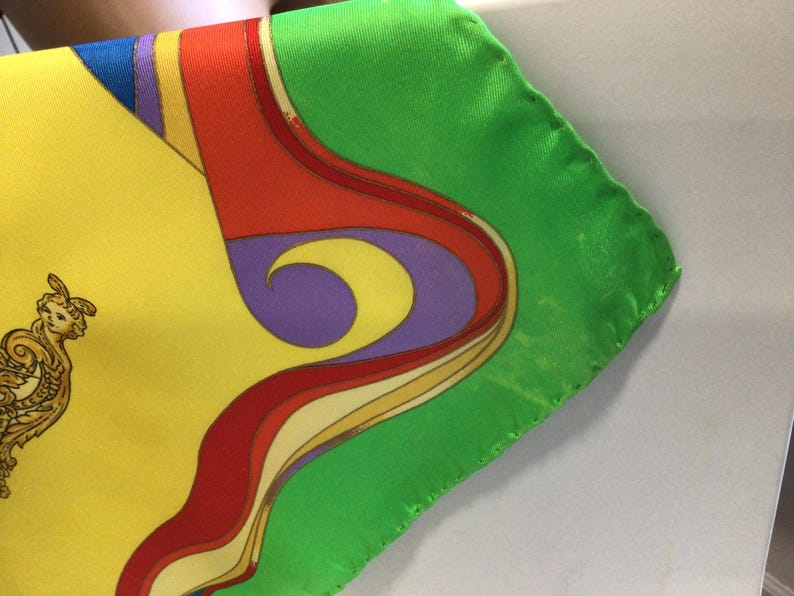 Puede incluir: Un pa&ntilde;uelo de seda colorido con un dise&ntilde;o vibrante. El pa&ntilde;uelo presenta un fondo amarillo con una figura dorada y negra adornada, y un borde verde. El dise&ntilde;o incluye formas curvas rojas, moradas y amarillas.