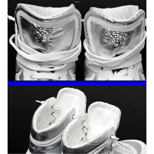 May include: High-top white sneakers with silver accents. The sneakers feature a silver panel on the back with a Medusa head emblem. White laces and a white interior complete the design. The shoes are displayed against a dark background.