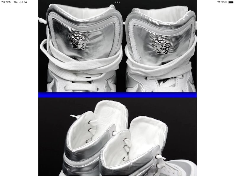May include: High-top white sneakers with silver accents. The sneakers feature a silver panel on the back with a Medusa head emblem. White laces and a white interior complete the design. The shoes are displayed against a dark background.