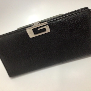 May include: Black leather wallet with a silver-tone metal clasp featuring a cut-out design. The wallet has a textured surface and a rectangular shape. The clasp is centered on the top edge.