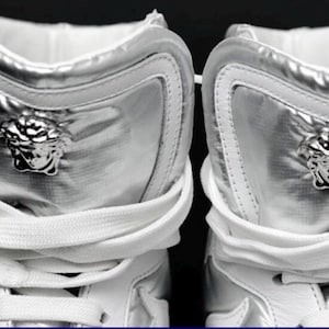 May include: Close-up of two silver high-top sneakers. The sneakers have a metallic sheen and feature a Medusa head emblem on the side. White laces and a white interior complete the design. The shoes are a luxury fashion item.