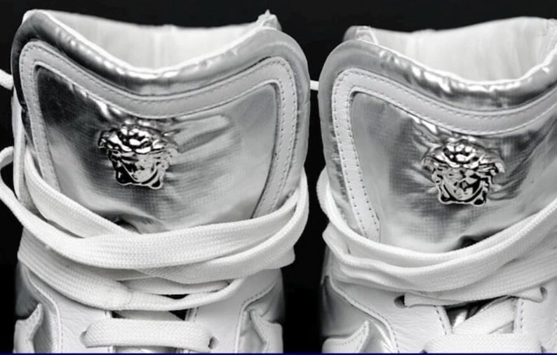 May include: Close-up of two silver high-top sneakers. The sneakers have a metallic sheen and feature a Medusa head emblem on the side. White laces and a white interior complete the design. The shoes are a luxury fashion item.
