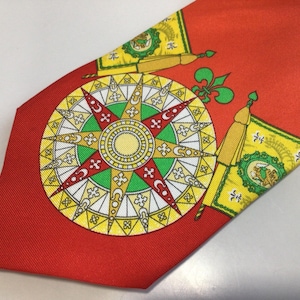 May include: Red silk tie with a detailed compass rose design in yellow, green, and white. The tie also features yellow and green flags with a crest and a fleur-de-lis. A classic accessory.