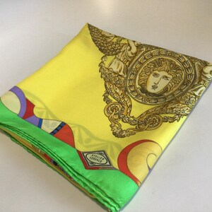 Puede incluir: Pa&ntilde;uelo de seda amarillo doblado con un borde verde. El pa&ntilde;uelo presenta un dise&ntilde;o de cabeza de Medusa dorada y el texto "ATELIER VERSACE". El borde tiene un patr&oacute;n geom&eacute;trico colorido.