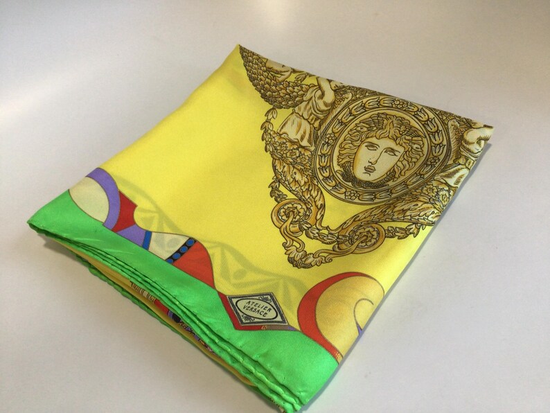 Puede incluir: Pa&ntilde;uelo de seda amarillo doblado con un borde verde. El pa&ntilde;uelo presenta un dise&ntilde;o de cabeza de Medusa dorada y el texto "ATELIER VERSACE". El borde tiene un patr&oacute;n geom&eacute;trico colorido.