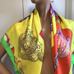 Puede incluir: Un pa&ntilde;uelo de seda amarillo con un dise&ntilde;o colorido. El pa&ntilde;uelo presenta un medall&oacute;n central con una cara y figuras, rodeado de detalles ornamentados. Los bordes est&aacute;n ribeteados con verde y otros colores. El pa&ntilde;uelo est&aacute; drapeado sobre un maniqu&iacute;.