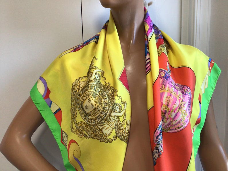 Puede incluir: Un pa&ntilde;uelo de seda amarillo con un dise&ntilde;o colorido. El pa&ntilde;uelo presenta un medall&oacute;n central con una cara y figuras, rodeado de detalles ornamentados. Los bordes est&aacute;n ribeteados con verde y otros colores. El pa&ntilde;uelo est&aacute; drapeado sobre un maniqu&iacute;.