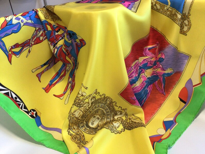Puede incluir: Un pa&ntilde;uelo de seda amarillo con figuras abstractas coloridas e im&aacute;genes cl&aacute;sicas. El pa&ntilde;uelo presenta un dise&ntilde;o vibrante con figuras rojas, azules y moradas, enmarcadas por un borde verde. El dise&ntilde;o incluye rostros cl&aacute;sicos y detalles ornamentados.