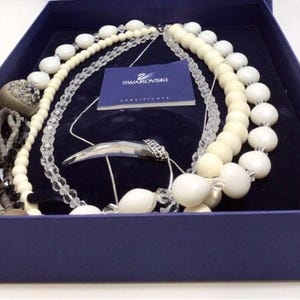 May include: A collection of necklaces in a navy blue box. The necklaces include white beaded strands, clear crystal strands, and a silver pendant. A small blue card with the Swarovski logo is also visible.